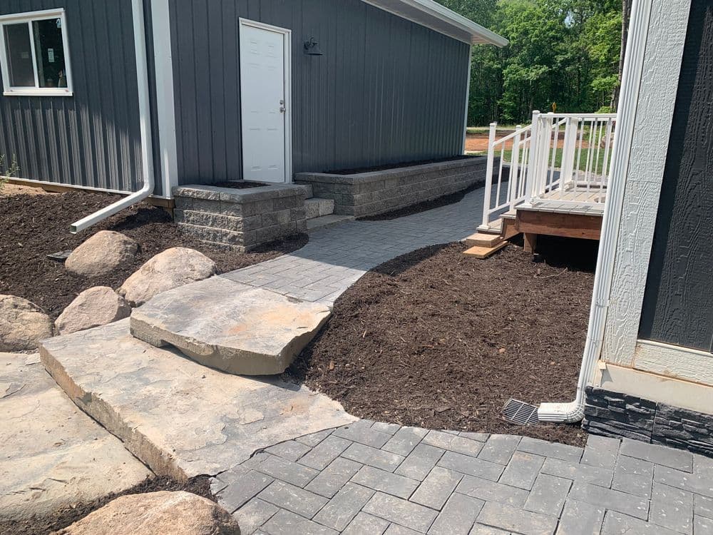 Paved walkway leading to a gray house with landscaping and stone features.