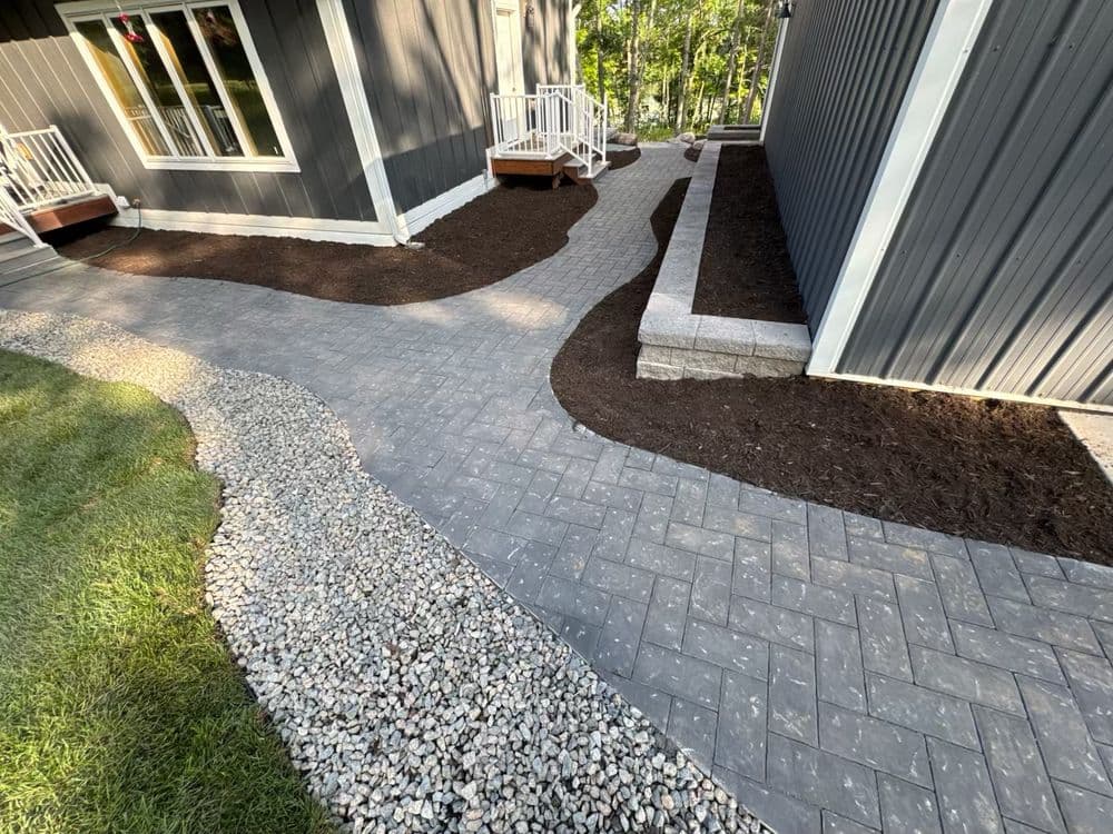 Paved walkway with gravel and mulch landscaping beside a modern home in a wooded area.