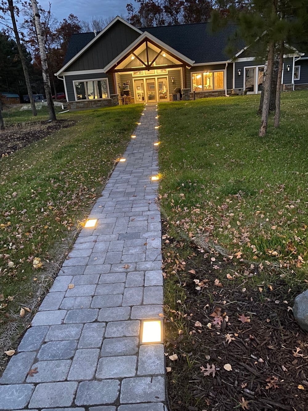 Illuminated stone pathway leading to a modern home at dusk, surrounded by grass and trees.