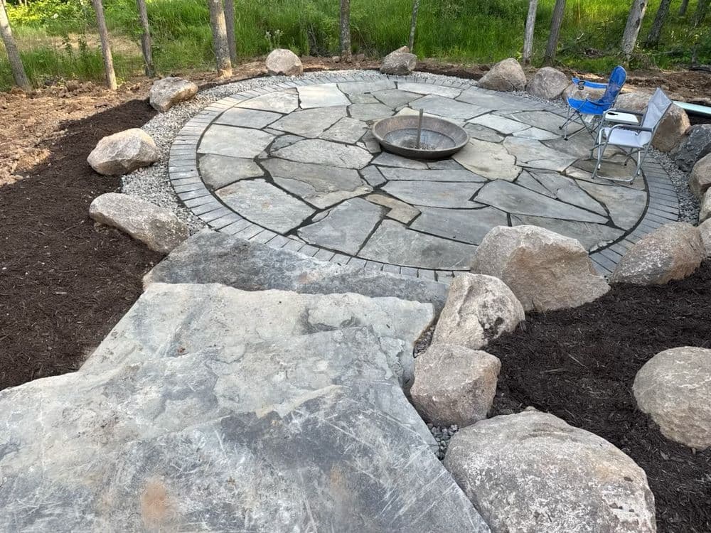 Stone fire pit area with large rocks and a blue chair, surrounded by gravel and mulch.