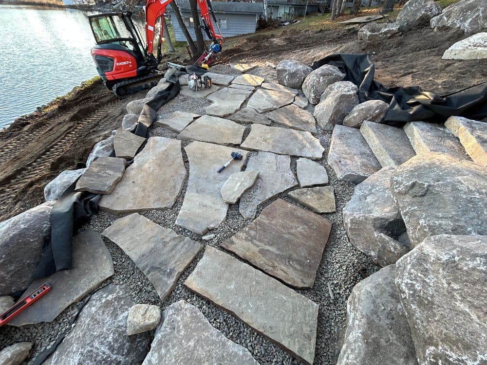 Natural stone pathway under construction with machinery and landscaping materials by a lake.