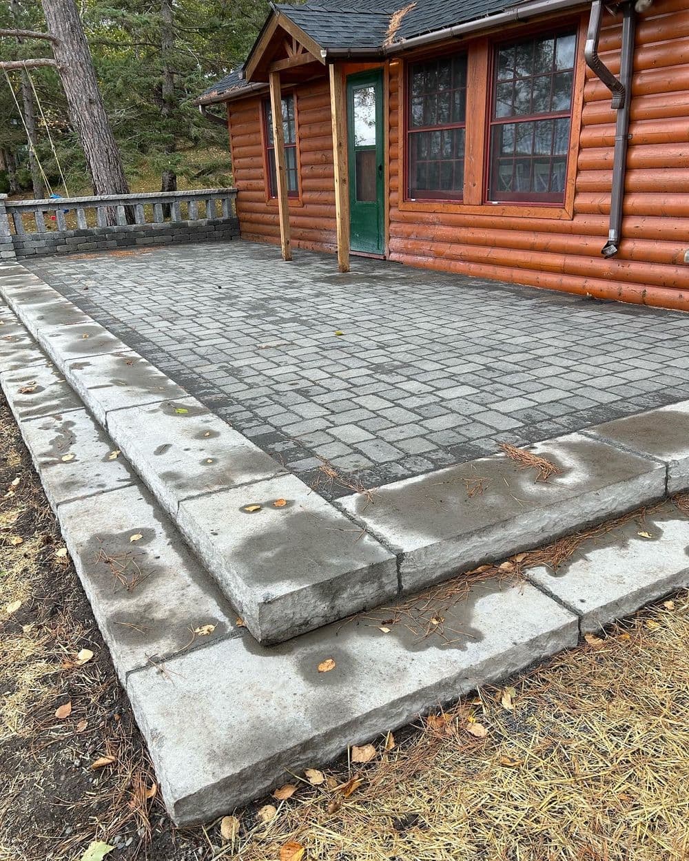 Log cabin exterior with new stone paver patio and steps, surrounded by trees.