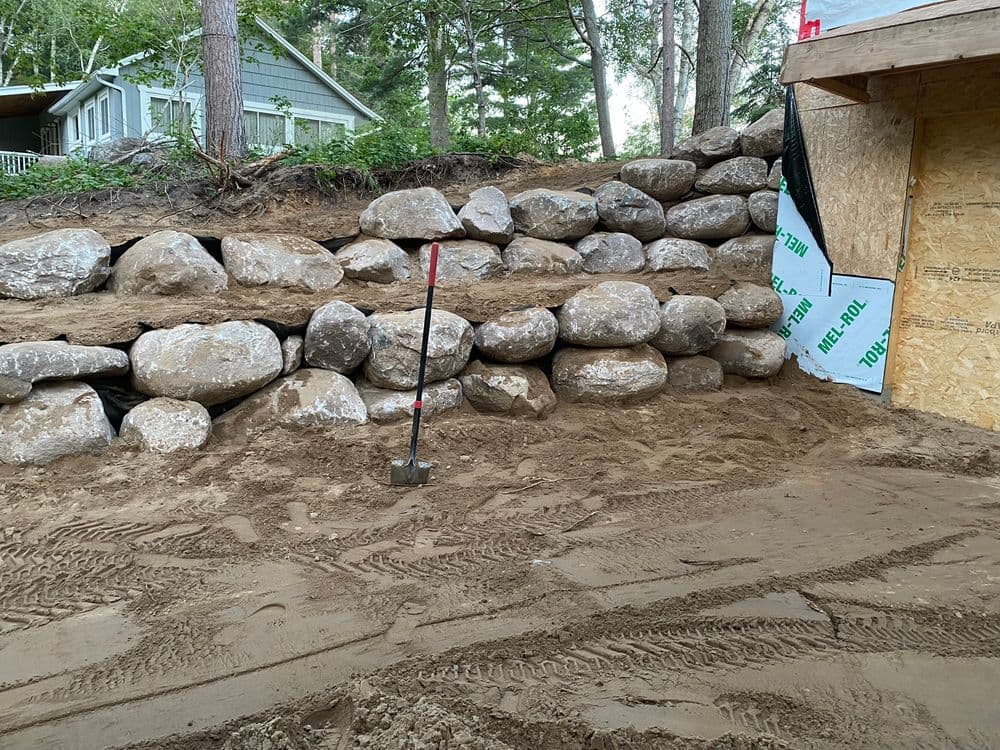 Rock retaining wall under construction with a shovel resting on sandy ground.