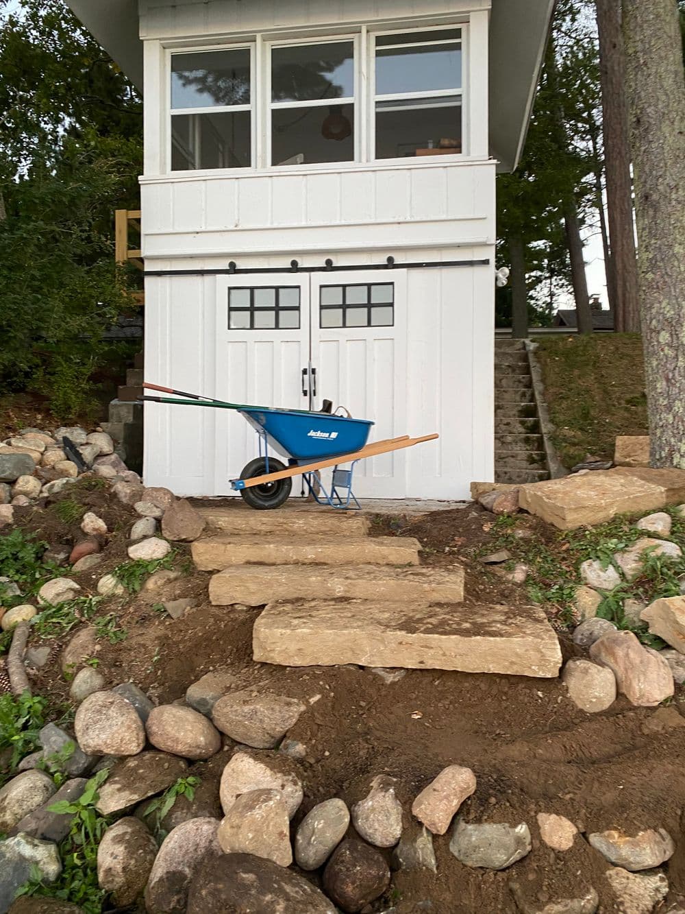 Blue wheelbarrow on stone steps leading to a white building surrounded by greenery.