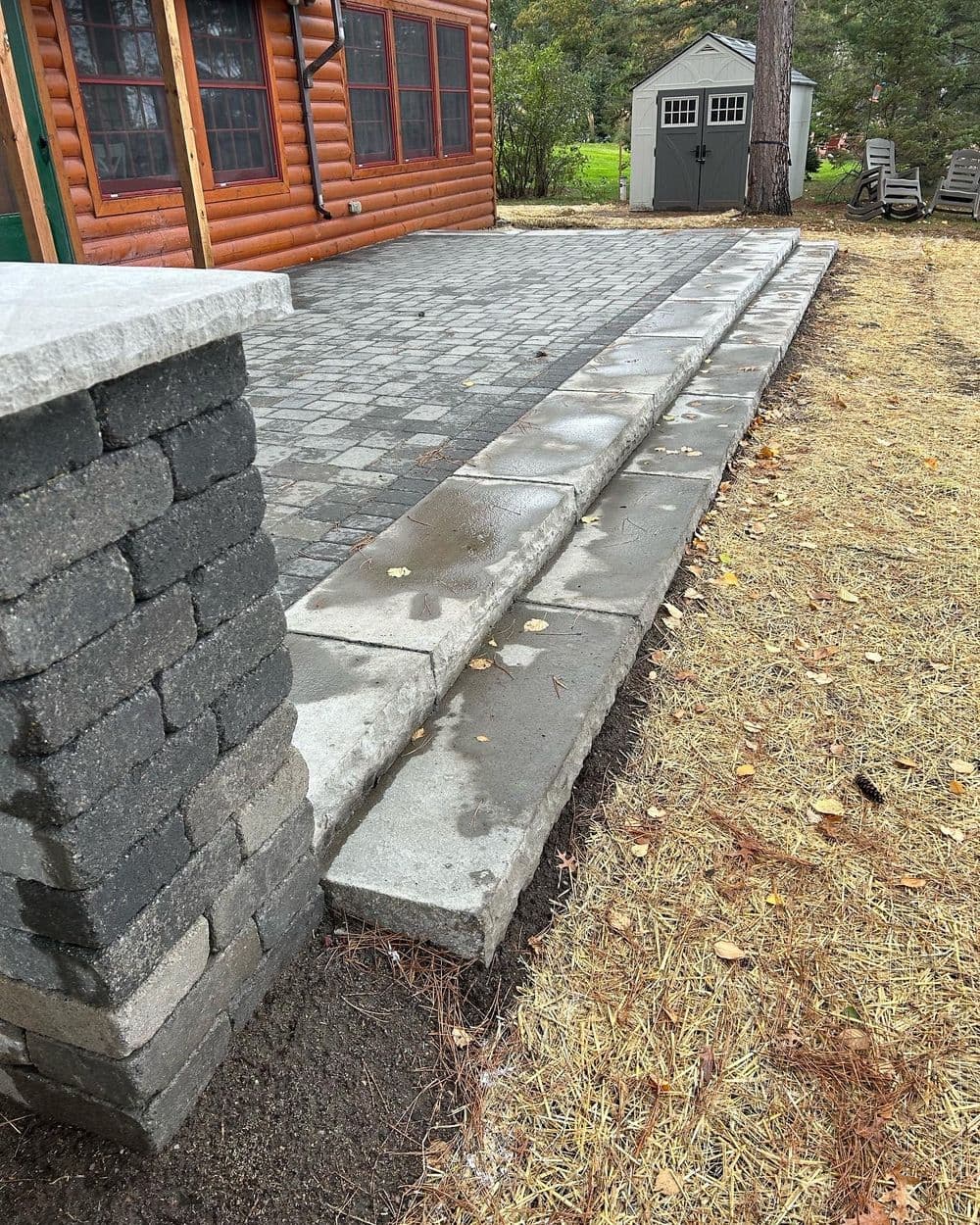 Paved stone steps leading to a log cabin patio with landscaping and a garden shed nearby.