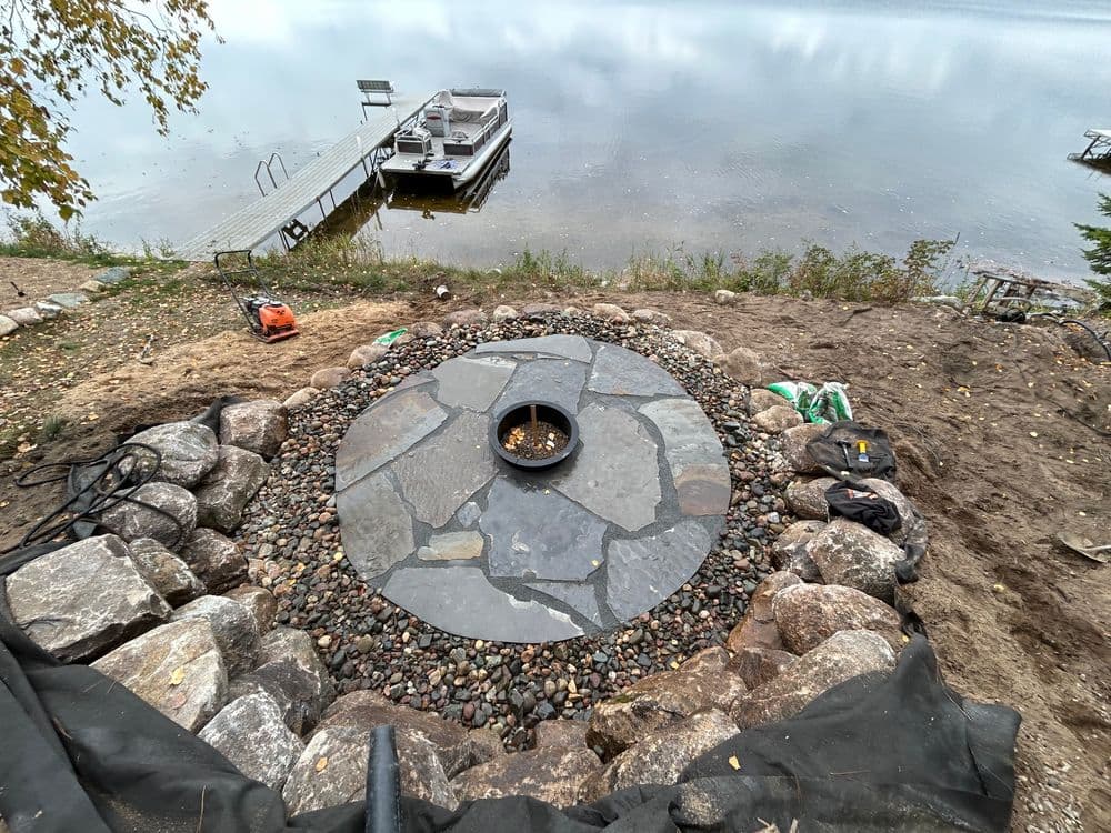 Stone patio with fire pit by a lake, surrounded by landscaping stones and tools.