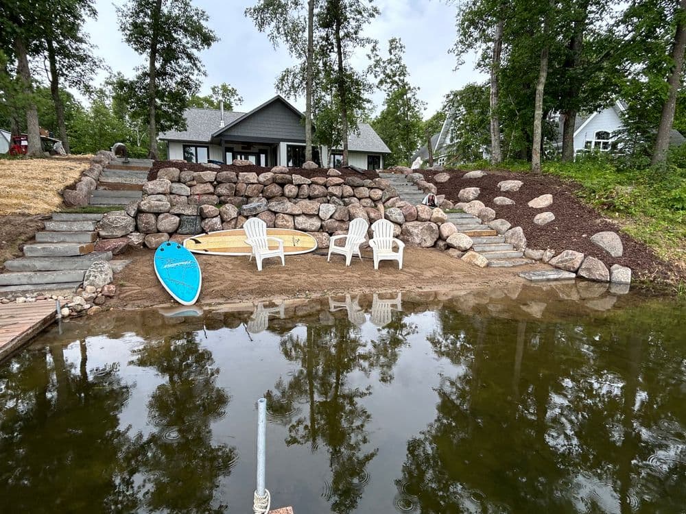 Lakeside retreat with chairs, paddleboards, and stone landscaping near calm water.