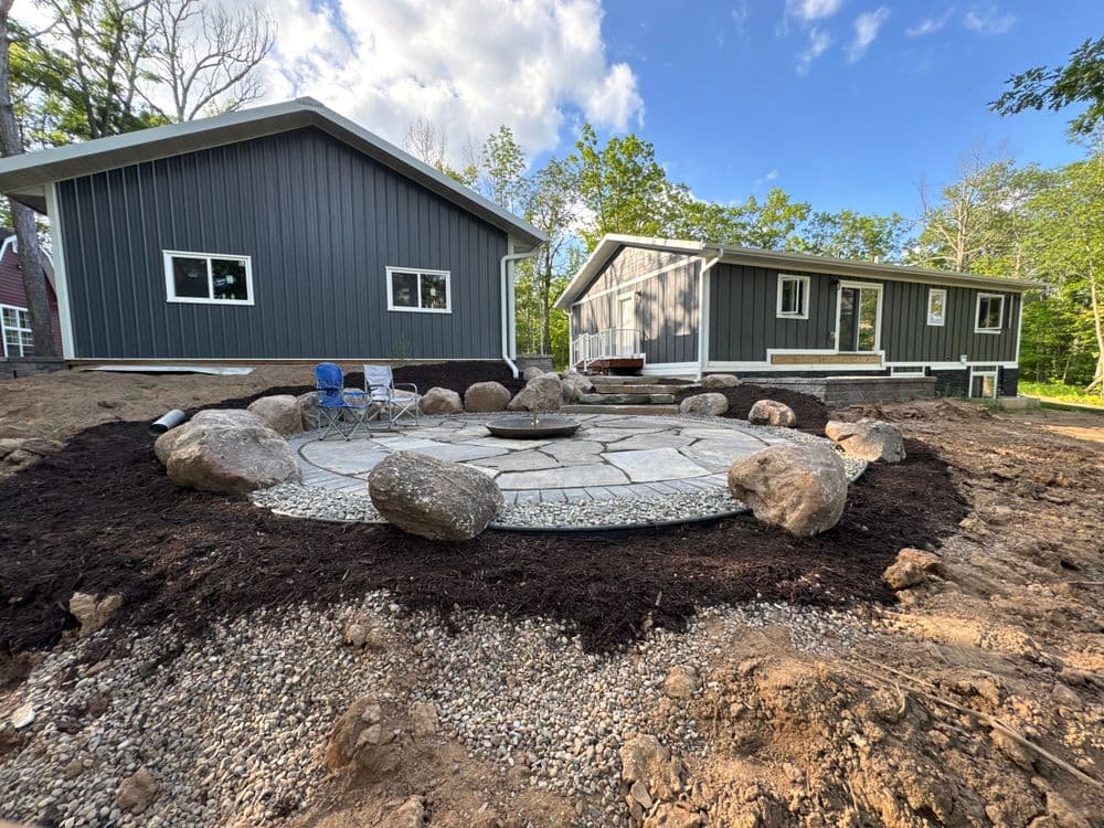 Modern backyard landscape with a stone fire pit, surrounded by boulders and mulch.