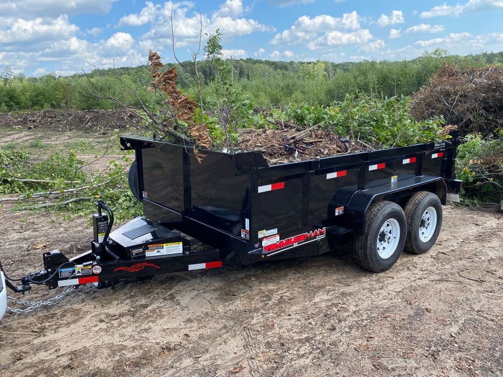 Black utility trailer loaded with branches and debris in a wooded area under a blue sky.