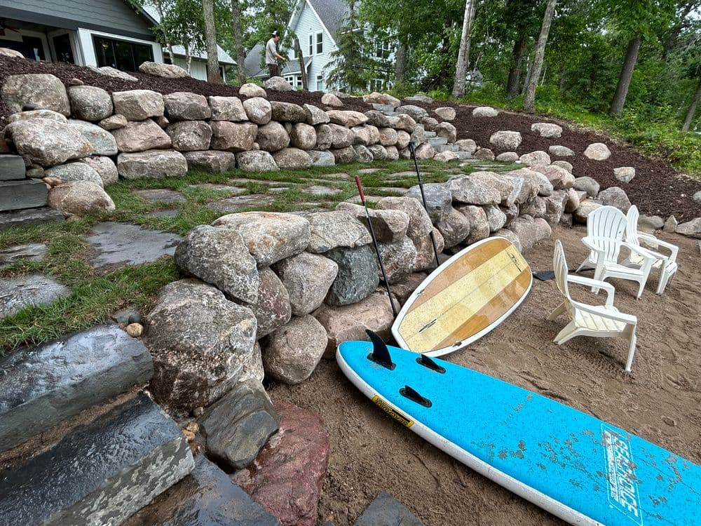 Surfboard and paddle resting on sandy shore, with stone retaining wall and chairs in background.