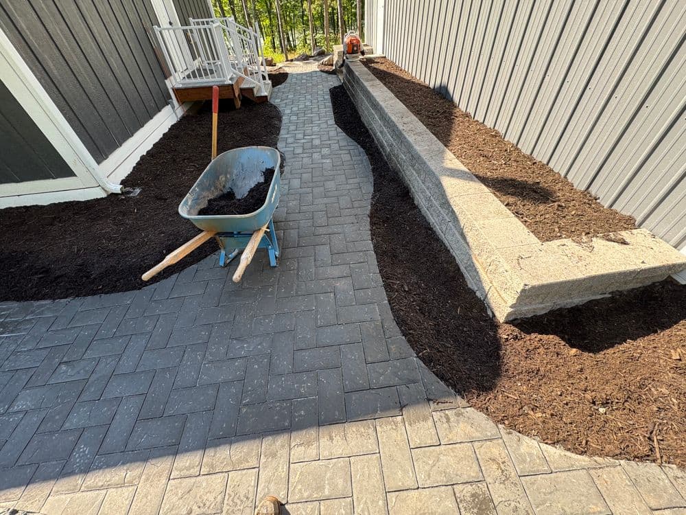 Paved walkway with mulch bed, wheelbarrow, and stone wall in a landscaped garden area.
