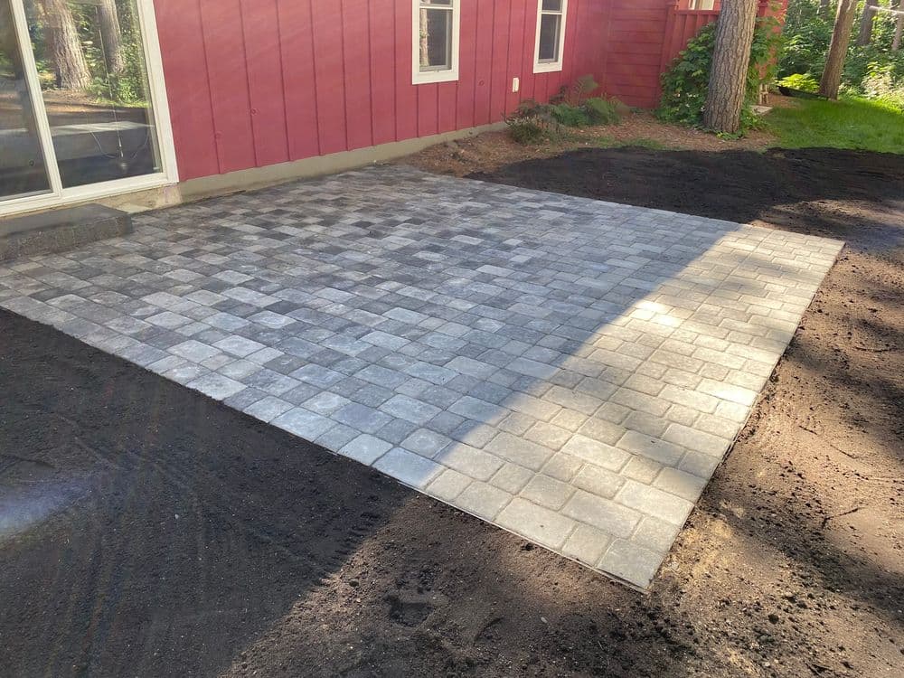 Newly installed gray paver patio next to a red house, surrounded by dirt and trees.