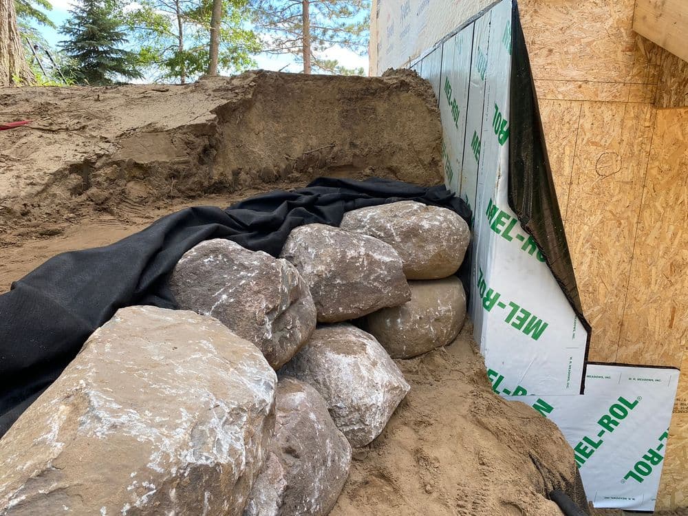 Rock wall construction with protective fabric and insulation materials in a landscaped area.