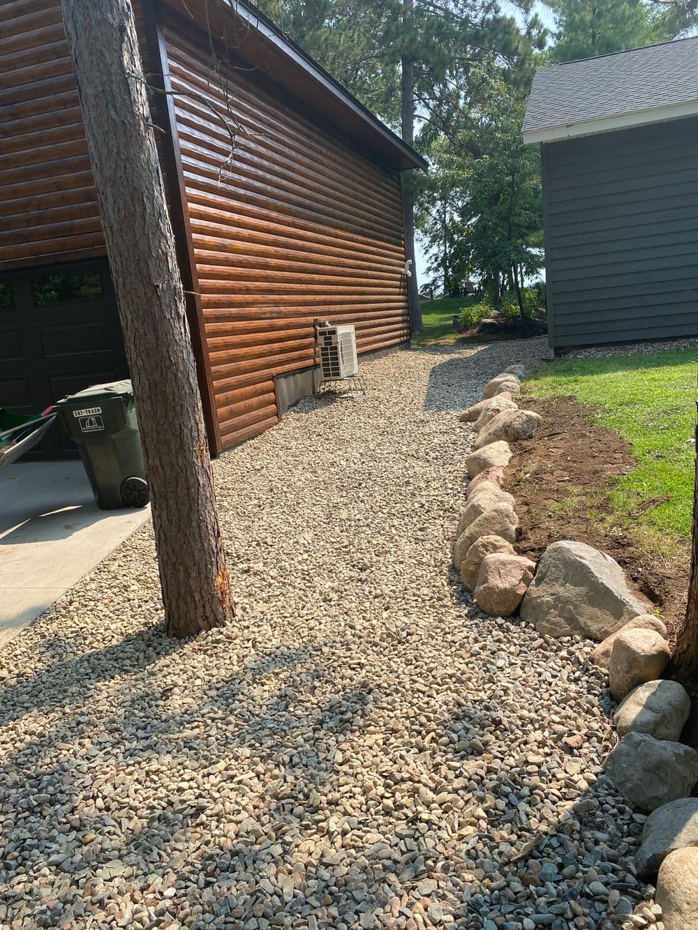 Gravel pathway alongside a wooden cabin and gray house, bordered by rocks and trees.