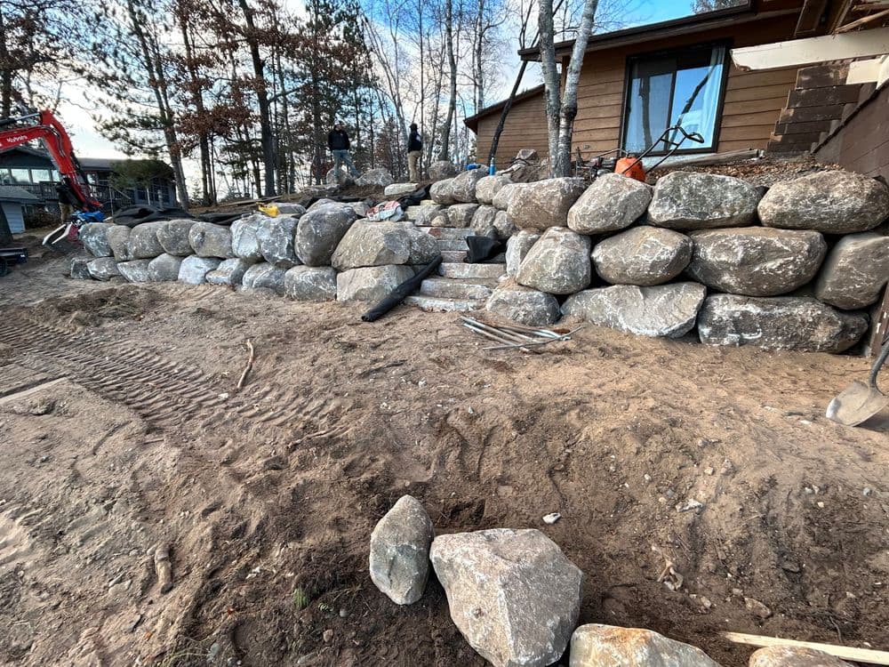 Landscaping project with large rocks forming a retaining wall beside a house under construction.