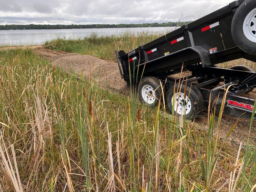 Dump trailer on gravel path near lake with tall grass and cloudy sky in background.