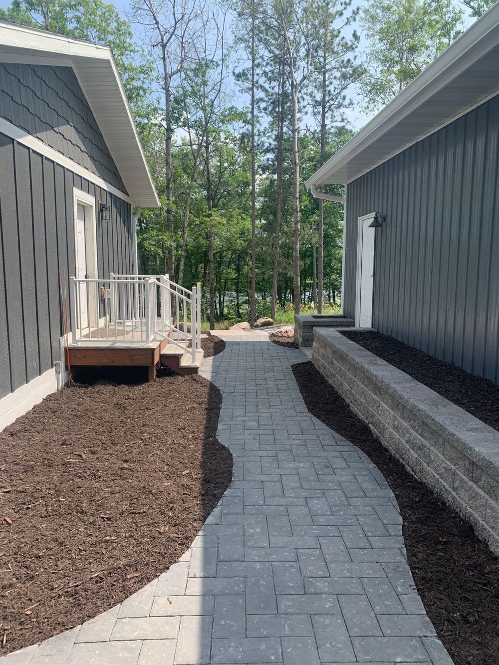 Paved walkway between modern homes, surrounded by mulch and trees in a sunny setting.
