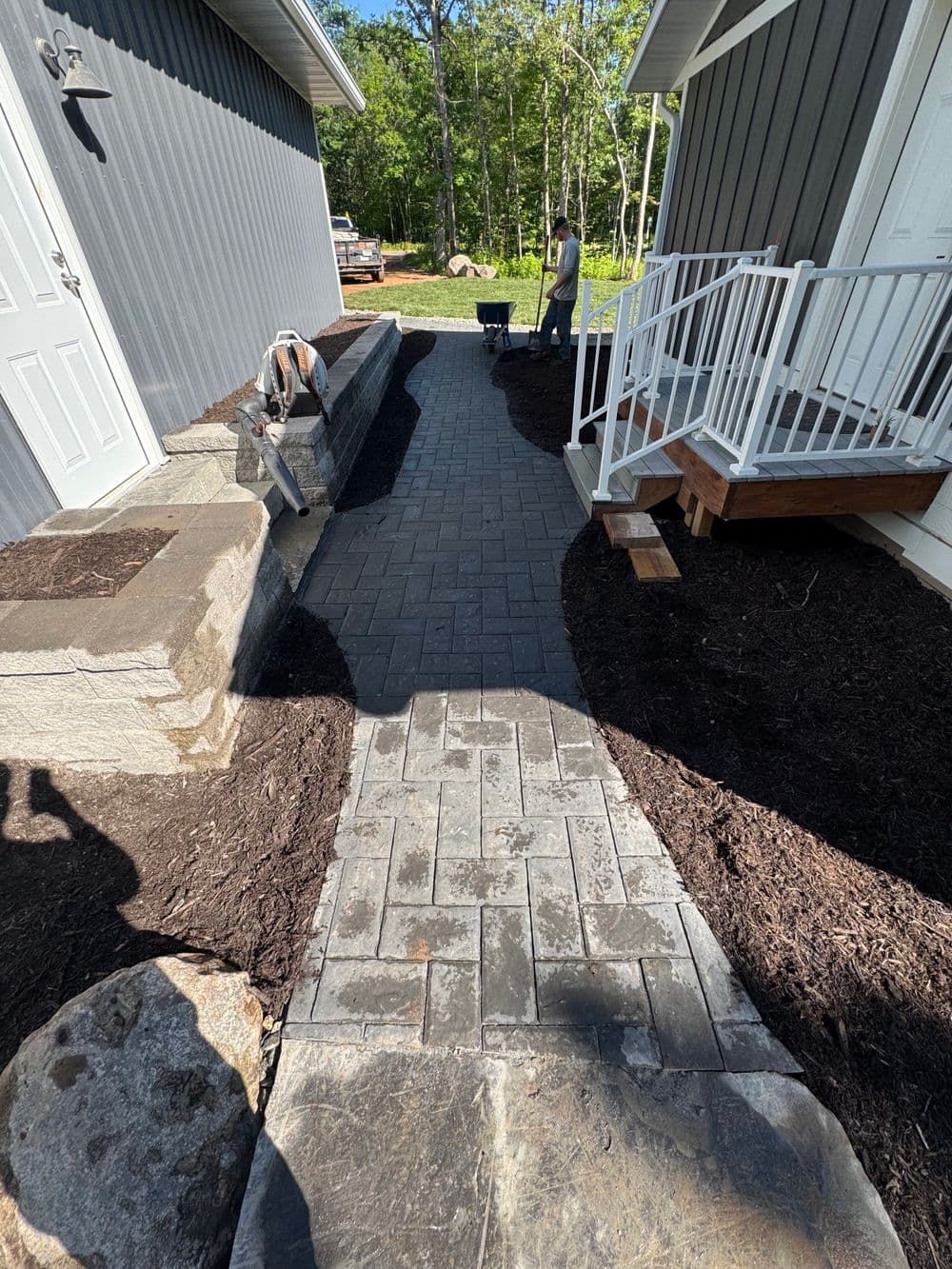 Paved walkway installation between two gray houses with landscaping in progress.