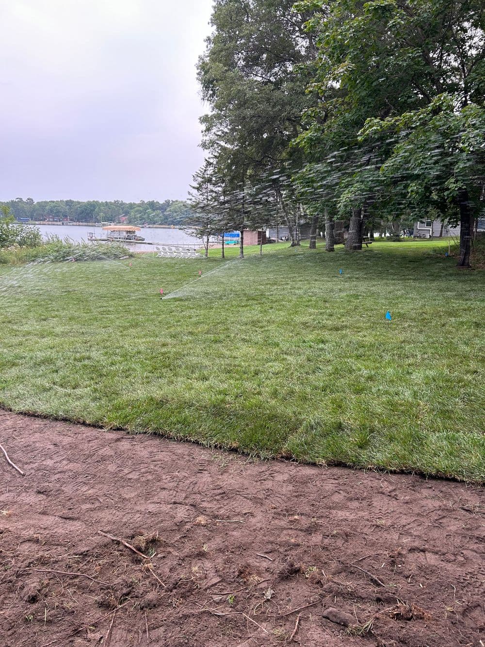 Lush green lawn with sprinklers near a lake, surrounded by trees and marked with flags.