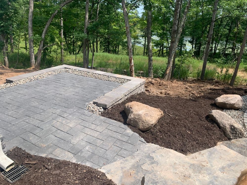 Backyard patio with paver stones, bordered by rocks and mulch, surrounded by trees and greenery.