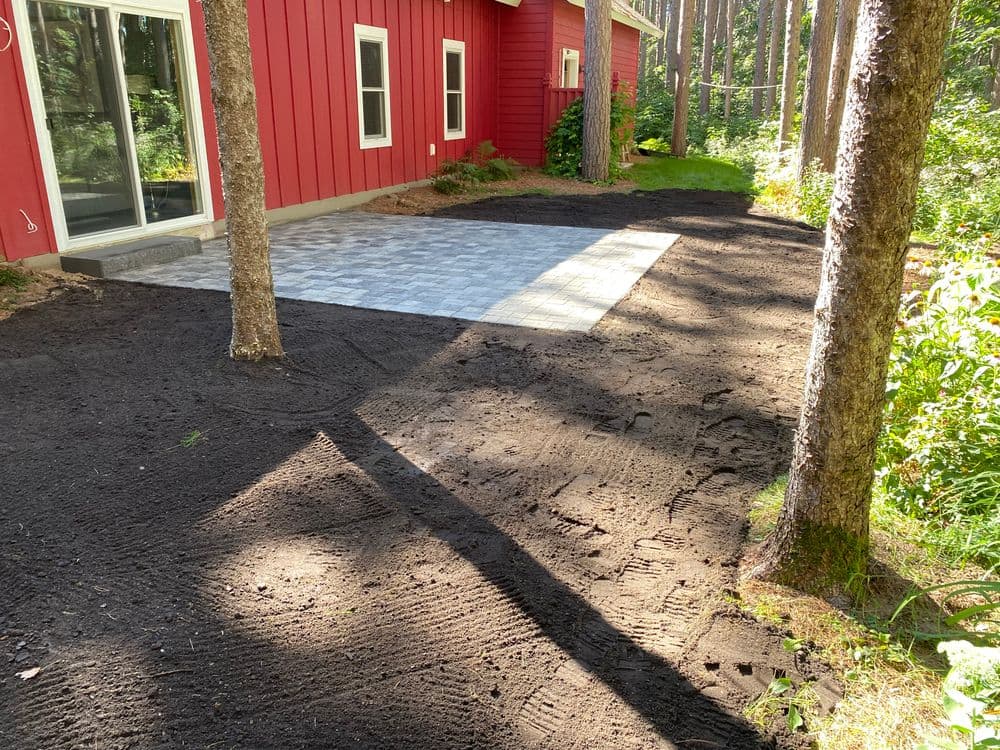 Paver patio installation next to a red house in a wooded area with fresh landscaping.