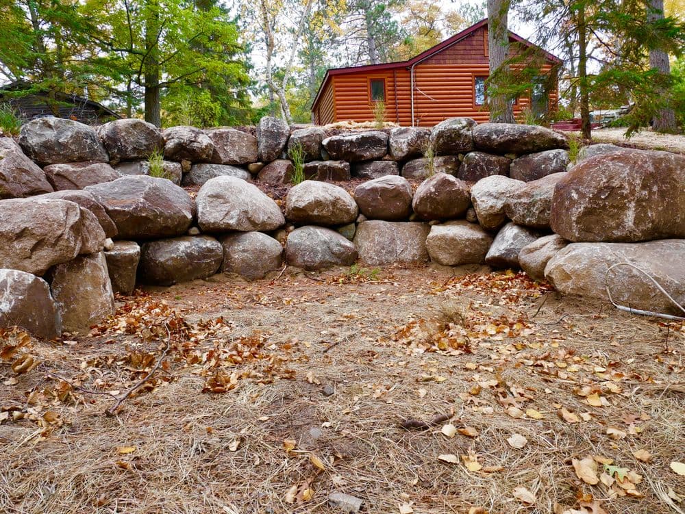 Stone retaining wall with autumn leaves and a log cabin in the background.