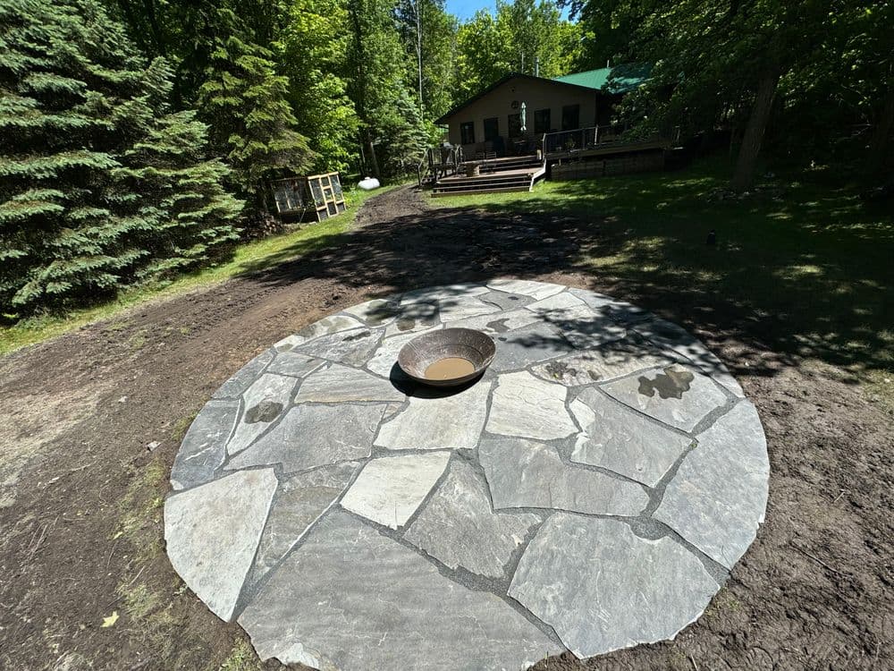 Stone patio with a circular fire pit in a forested backyard with a cabin in the background.