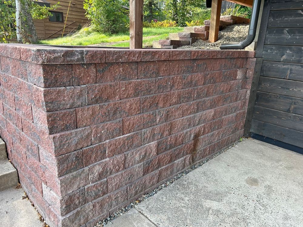 Red stone retaining wall beside steps and a wooden building, enhancing outdoor landscaping.