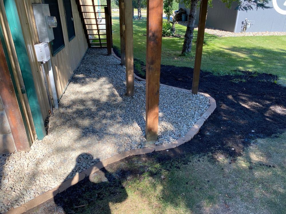 Gravel pathway and landscaping under a porch with wooden supports and a black mulch border.