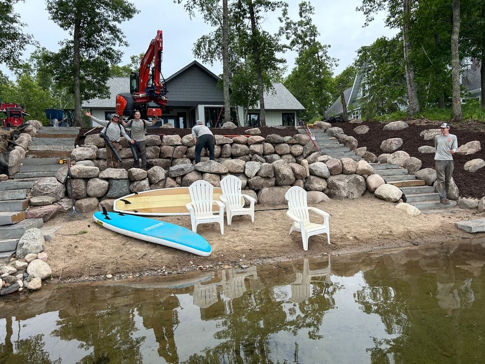 Construction crew landscaping a lakeside with rocks, paddleboards, and Adirondack chairs.