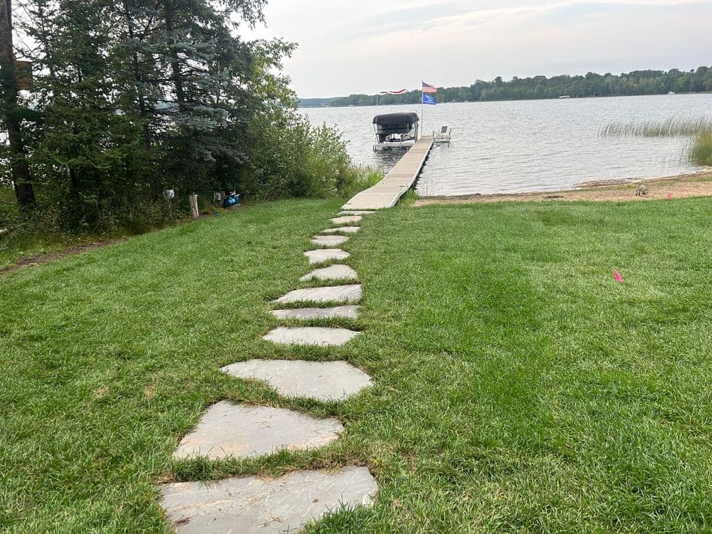 Stone pathway leading to a lake dock surrounded by green grass and trees.