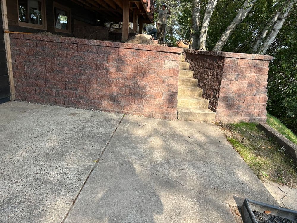 Stone wall with stairs leading to a raised area beside a house and trees in the background.