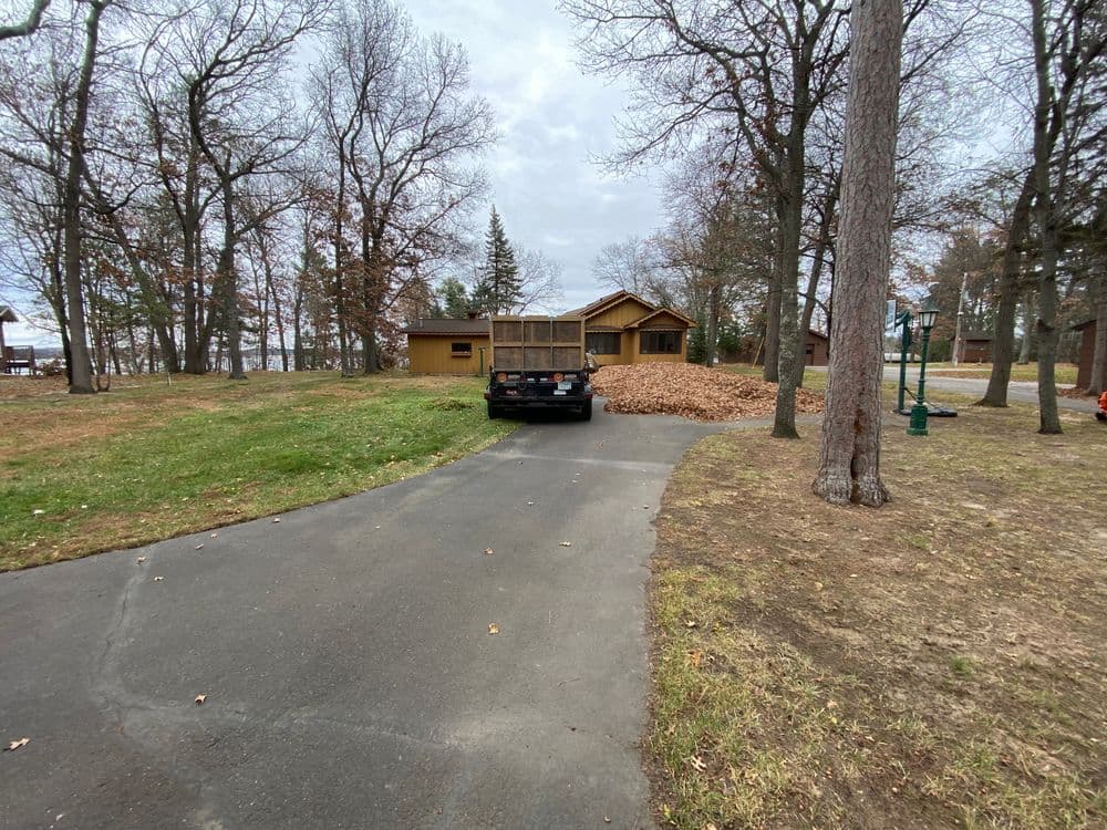 Black truck parked on driveway with piles of autumn leaves near a wooden house in a forested area.