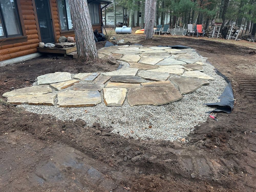 Flagstone pathway installation with gravel base in a landscaped yard near a cabin.