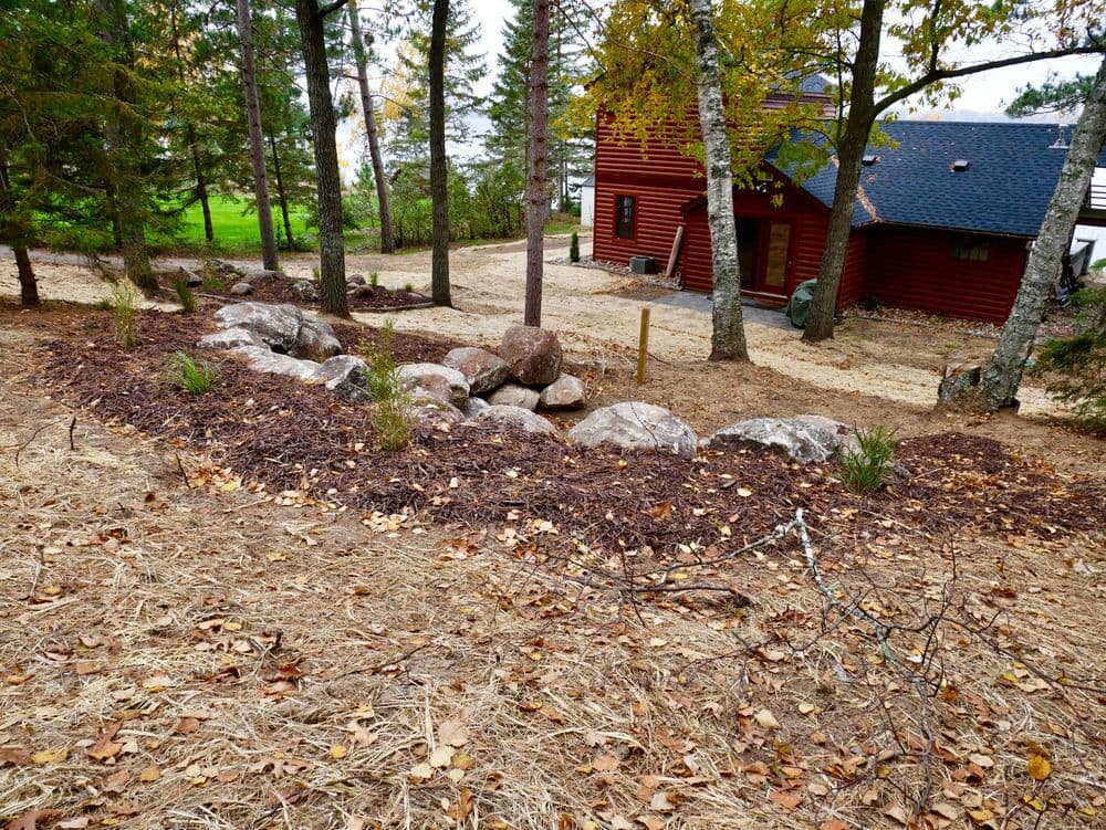 Landscaped garden with rocks and mulch near a red wooden house surrounded by trees.