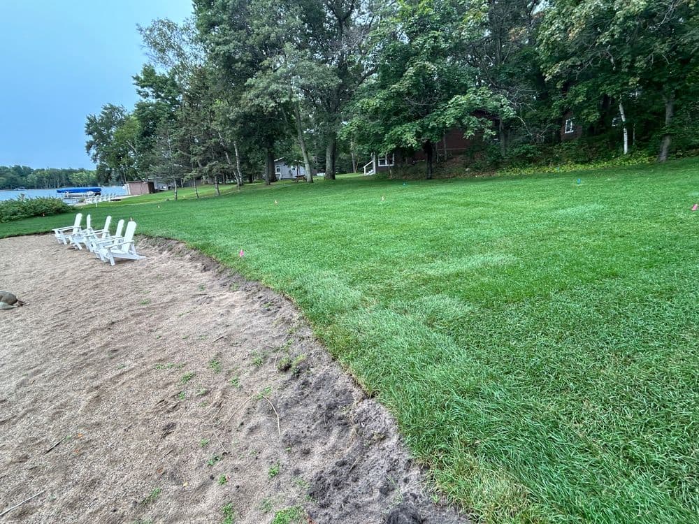 Lush green lawn with white chairs and sand area, surrounded by trees and a lake in background.