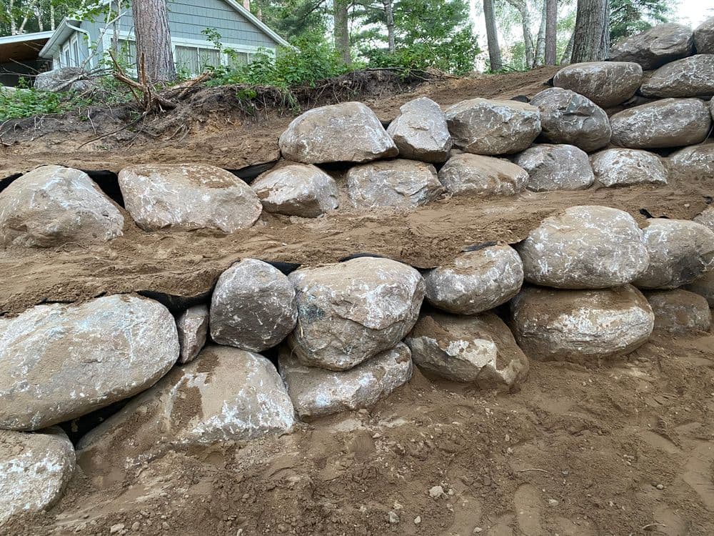 Stone retaining wall construction with large boulders and sandy soil background in a wooded area.