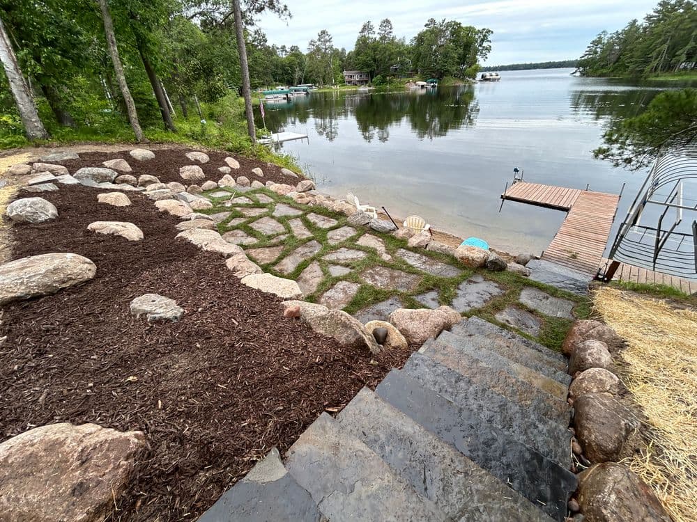 Lakeside stone pathway with landscaped rocks and wood dock by a tranquil lake.