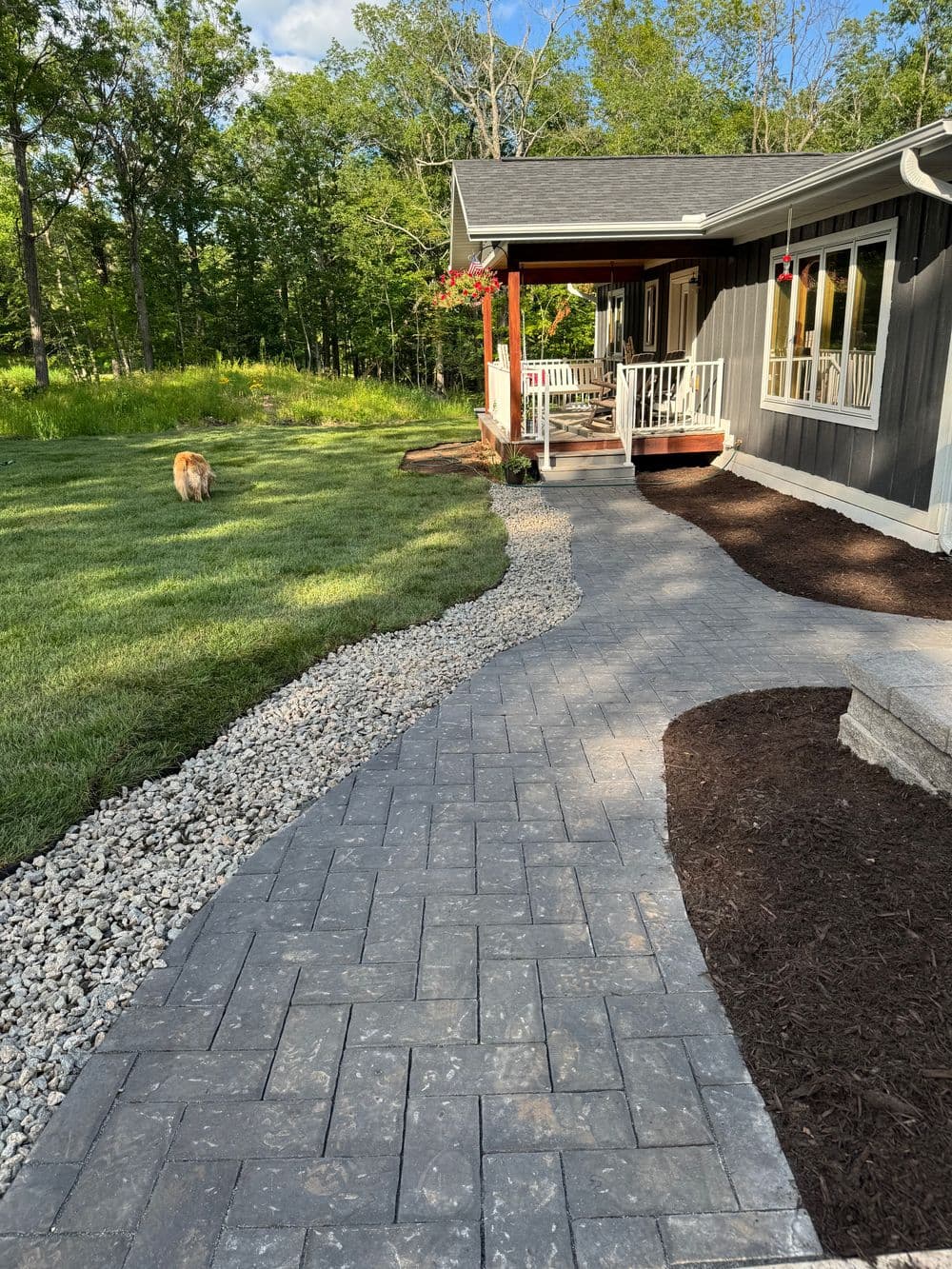 Paved walkway leading to a house with a dog nearby and lush greenery in the background.
