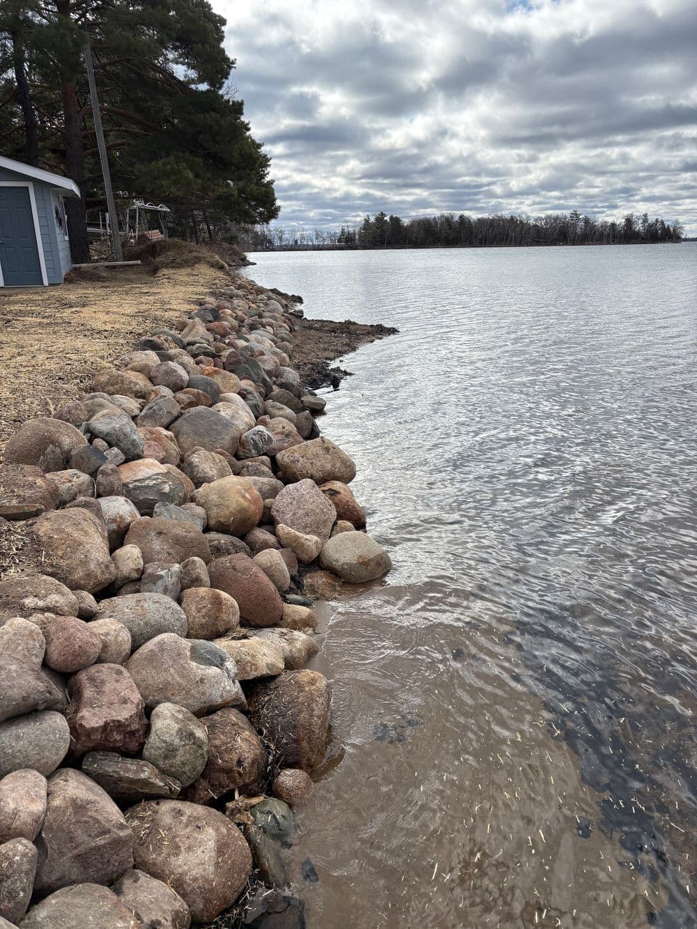 Rocky shoreline with calm water and cloudy sky at a lakeside in a rural setting.