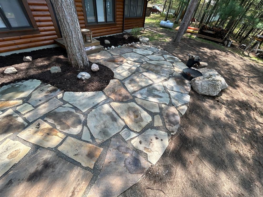 Natural stone patio installation with wooden cabin in background, surrounded by landscaping.