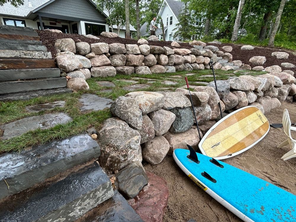 Stone wall landscaping with paddleboards near sandy beach and steps leading to a house.