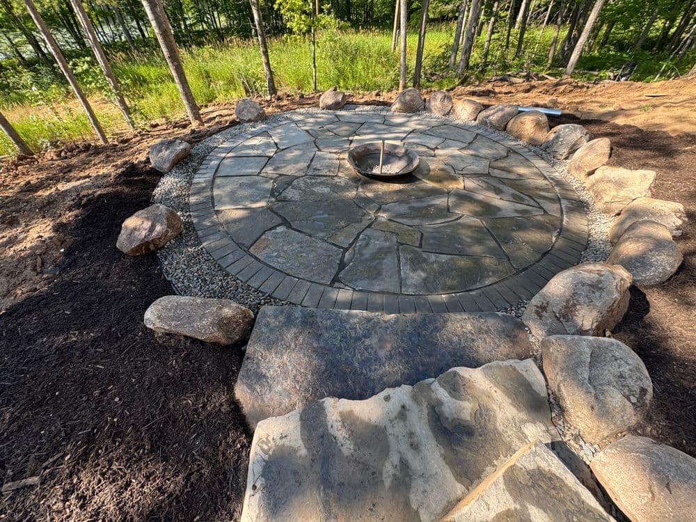 Stone circular patio with a fire pit, surrounded by natural landscaping and large rocks.