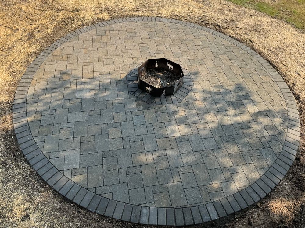 Circular stone patio with a fire pit in the center, surrounded by grass and tree shadows.